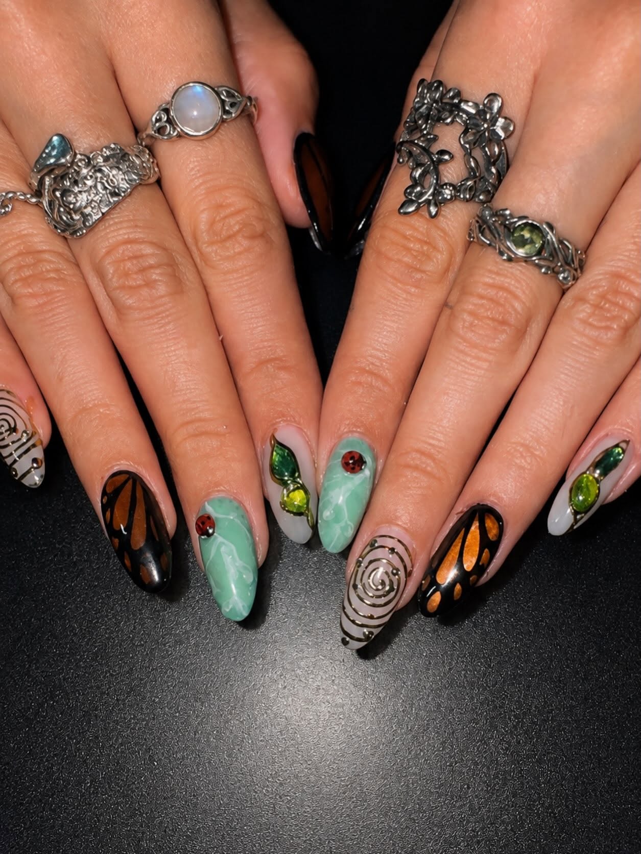 Long almond nails with hand-painted monarch butterfly wings, mint marbled nails with ladybirds, gold spirals and green gemstone leaf charms.