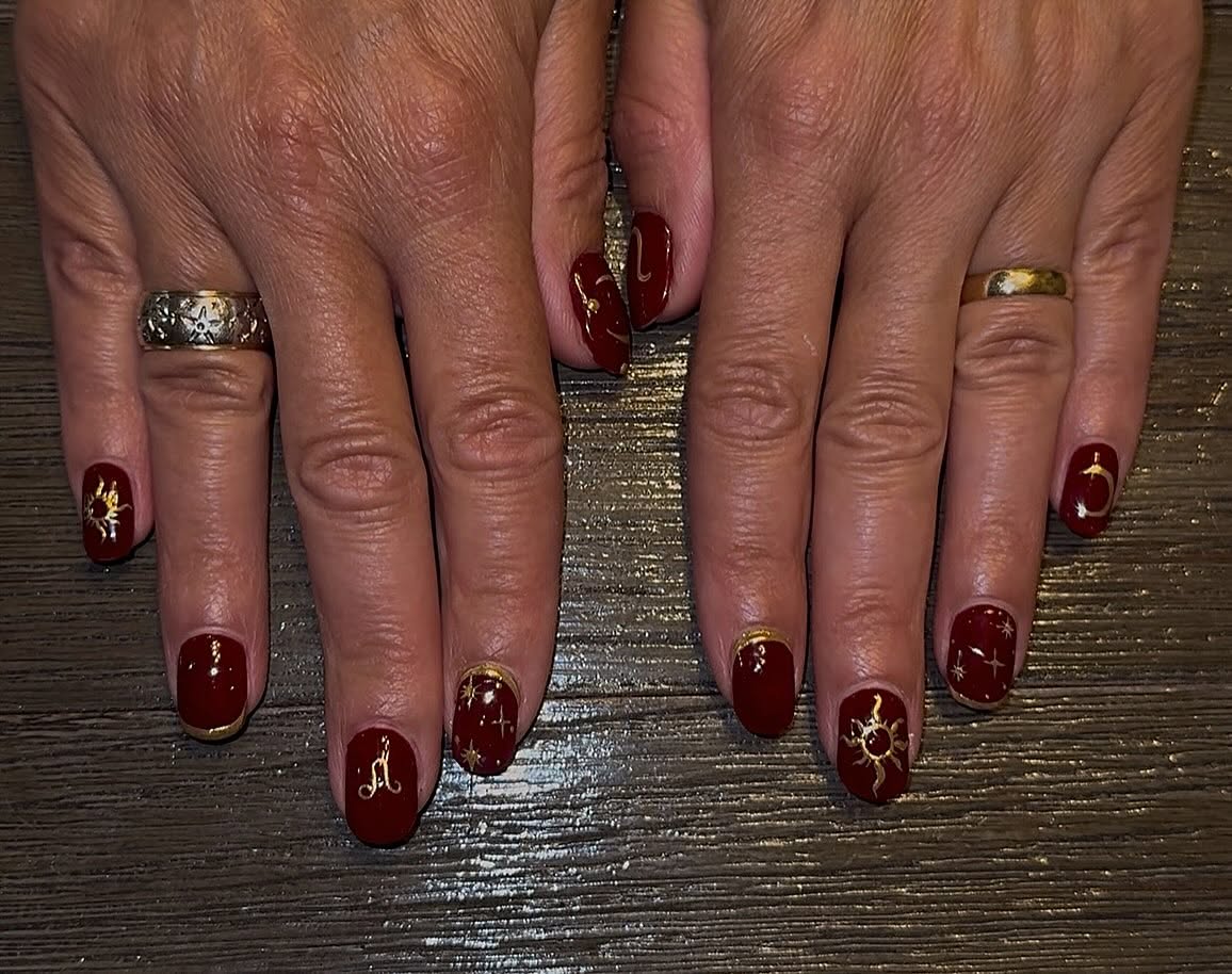 Short square nails in deep red with hand-painted gold suns, crescent moons, stars and a zodiac glyph.