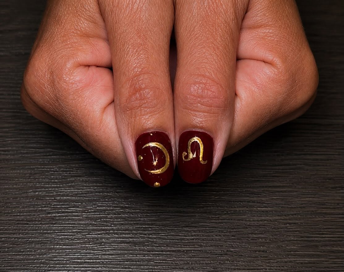 Close-up of two deep red nails, one with a gold crescent moon and star, the other with a gold Leo zodiac glyph.
