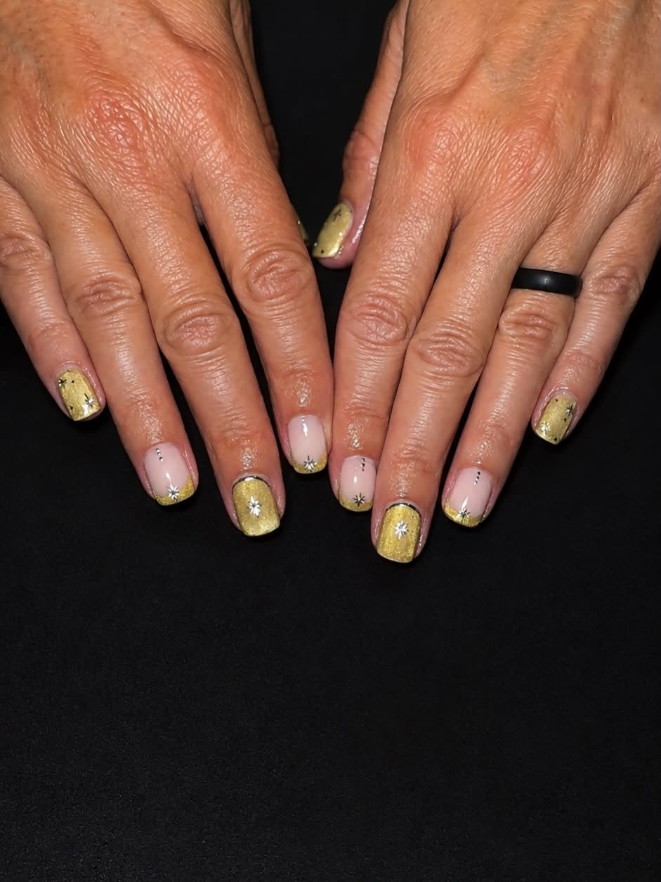 Short square gel nails in gold shimmer with a nude base and small hand-painted white starbursts and dots.