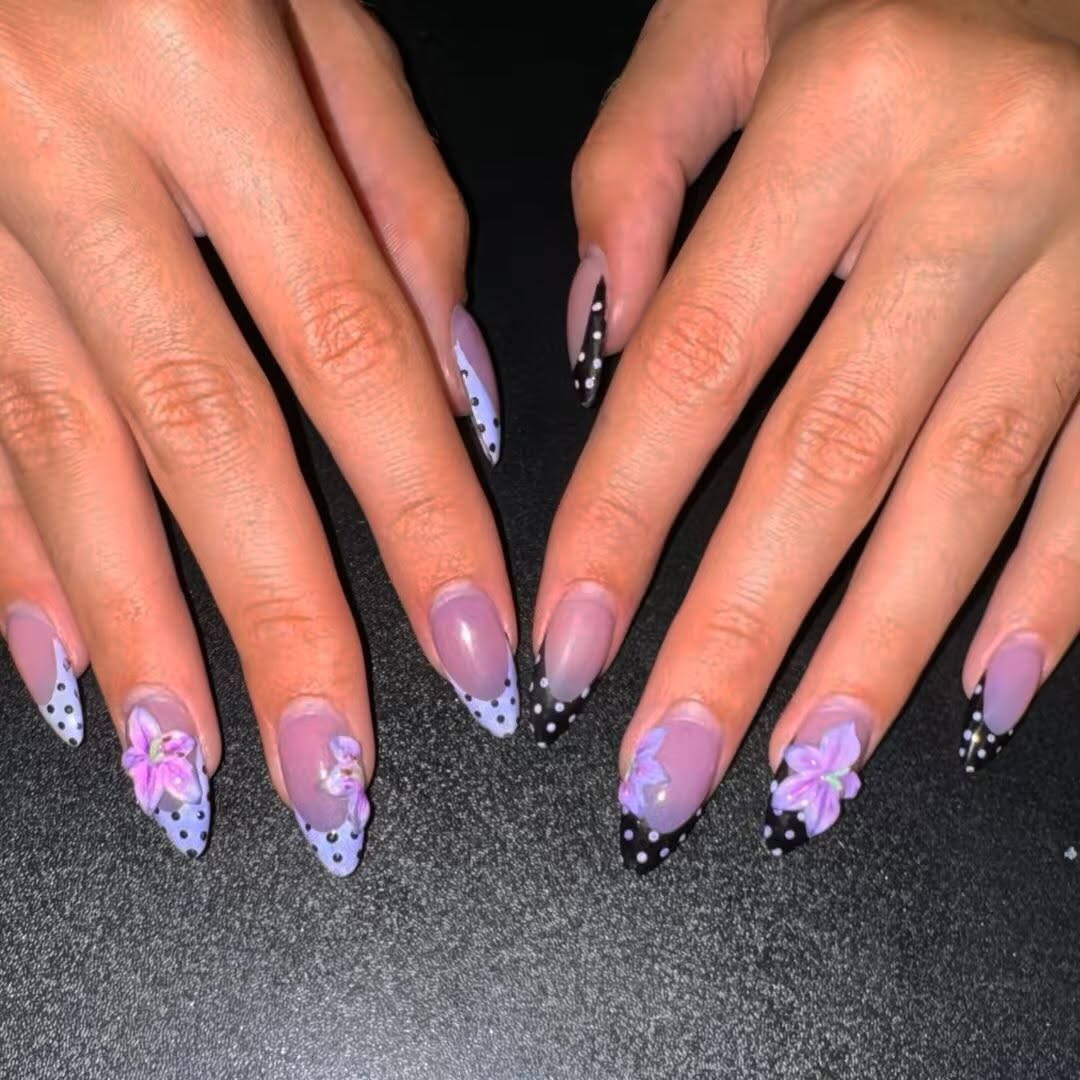 Long almond Gel-X nails with black and white polka-dot French tips over a lilac base, finished with raised 3D purple flowers.